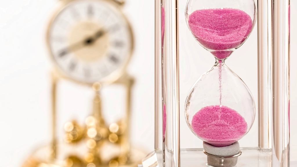 A glass hourglass with pink sand running through it, in front of a blurred gold mantel clock.