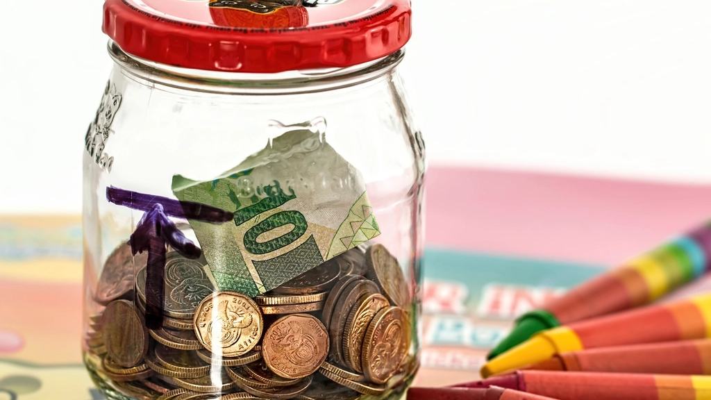 A glass savings jar with a red lid holding coins and a folded banknote, next to a row of crayons.