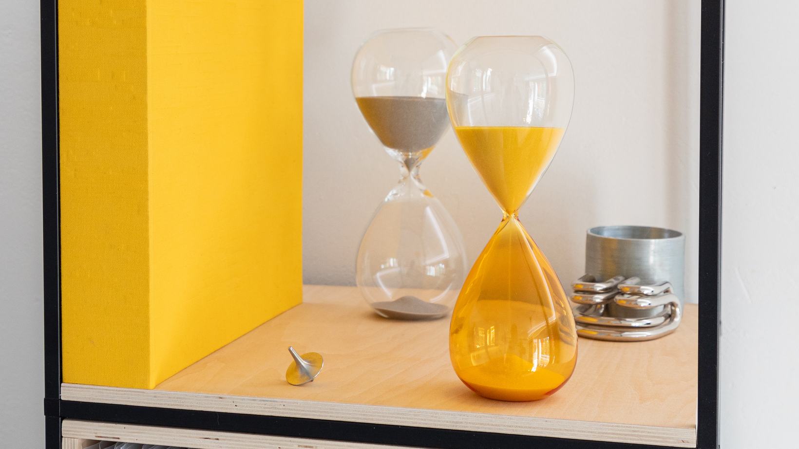 Two hourglasses on a shelf, one with grey sand and one with orange sand running out
